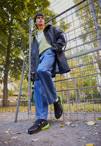 Svarta sportskor med gröna detaljer, ljusgrön t-shirt med en logotyp, kombinerat med lösa blå jeans och en svart regnjacka.