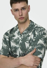 Cămașă cu mânecă scurtă, cu butoni, având un model cu frunze tropicale verzi pe un fundal alb, realizată dintr-un material ușor și neted.