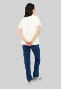 Creme korte mouwen t-shirt met een klein rood logo op de achterkant, gecombineerd met blauwe spijkerbroek en grijze sneakers. Achtergrond is effen grijs.