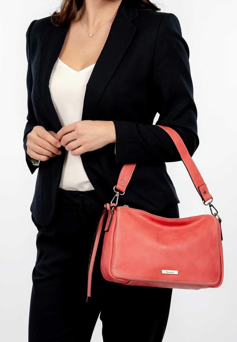 Femme portant un blazer noir et un haut blanc, tenant un sac à main en cuir corail avec des ferrures argentées suspendu à son bras.
