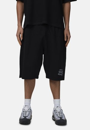 Osoba ubrana w luźne czarne szorty z białym napisem na lewej nodze, białe skarpety do kostki, szaro-białe sneakersy oraz czarną koszulkę na jednolitym tle.