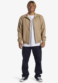 Tan lightweight jacket with a stand-up collar, zip closure, and two side pockets, worn over a white t-shirt, paired with dark blue jeans and black sneakers.