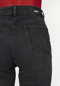 Primer plano de una persona que lleva unos pantalones de mezclilla negros con bolsillos traseros y una etiqueta negra que dice "JJXX" en la cinturilla.