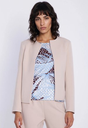 Kobieta ubrana w beżową marynarkę i spodnie, nałożone na wzorzysty top z abstrakcyjnym kwiatowym wzorem w kolorach niebieskim, białym i ciemnobrązowym, stojąca na jednolitym tle.