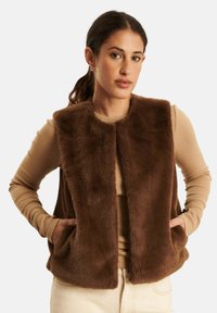 Brązowa kamizelka z sztucznego futra z okrągłym dekoltem, otwartym przodem i bocznymi kieszeniami, noszona na jasnobrązowej bluzce z długim rękawem.