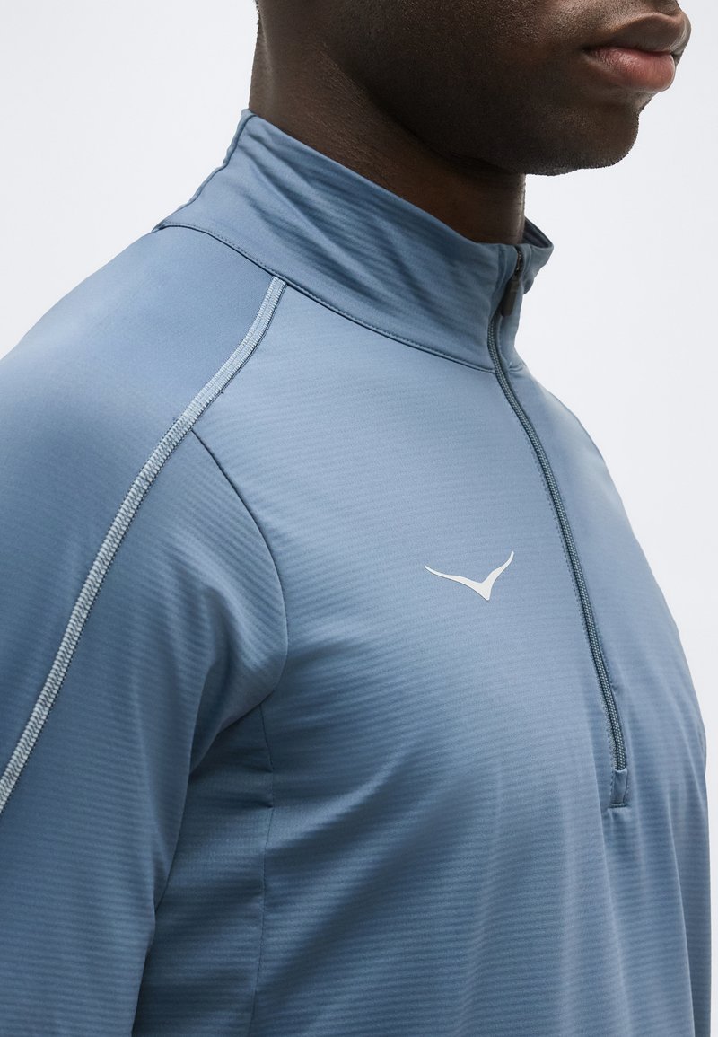 Primer plano de una persona que lleva una camiseta deportiva azul con cremallera parcial y un pequeño logo blanco en el pecho, con detalles de costuras visibles.