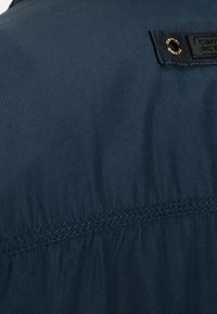 Ciemnoniebieska tkanina z poziomym przeszyciem i małą czarną prostokątną naszywką przymocowaną metalowym pierścieniem w górnym prawym rogu.