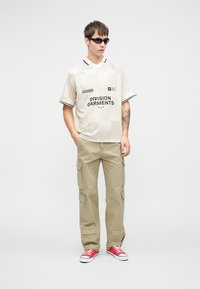 Beige t-shirt med kort ærme med sorte detaljer og logo, sammen med khaki cargobukser med lommer og røde sneakers.