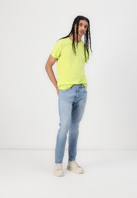 Neon zöld, rövid ujjú póló világoskék, slim-fit farmernadrággal és krémszínű sneakerrel párosítva, fonott hajjal, leengedve.