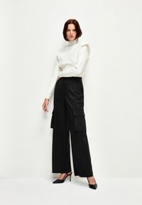 Weißer Pullover mit hohem Kragen und strukturierten Details, kombiniert mit schwarzen breiten Cargo-Hosen mit Nadelstreifen und schwarzen spitzen Pumps.