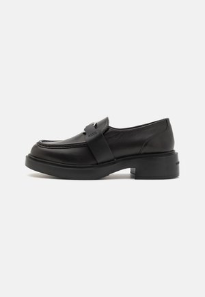 Mocassin en cuir noir avec une semelle épaisse, un petit talon bloc et un large détail de sangle sur le dessus, sur un fond blanc.