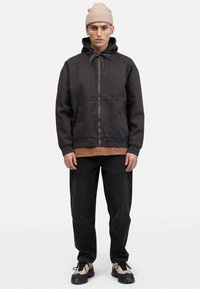 Black hooded jacket with a zipper, two side pockets, and a drawstring, layered over a brown shirt. Paired with black pants and beige sneakers.