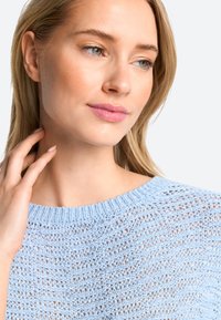Lichtblauwe gebreide trui met een losse, open structuur en een ronde halslijn, gepresenteerd op een model met neutrale make-up en licht haar.