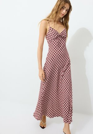 Jeune femme portant une longue robe rouge et blanche à carreaux sans manches avec de fines bretelles, debout devant un fond blanc uni.