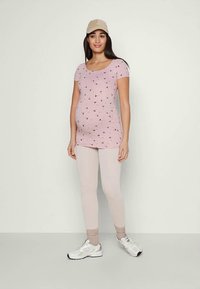 Maternity top in light pink with black floral print, paired with light beige leggings and white athletic shoes. Cap and sporty design visible.
