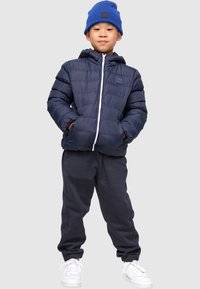 Marineblå sørvest med hette og hvit glidelås, kombinert med mørkegrå joggebukser og hvite joggesko. Blå strikket lue på hodet.