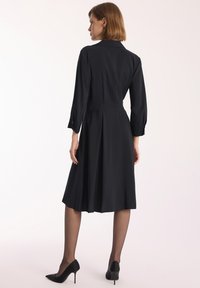 Czarna sukienka z gładkiego materiału, z podkreśloną talią, plisowanym dołem i rękawami o długości trzech ćwierci. Noszona z czarnymi szpilkami.