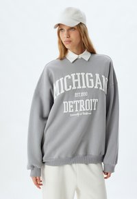 Grauer, übergroßer Kapuzenpullover mit weißer "MICHIGAN DETROIT" Schrift und geripptem Saum, über einem weißen Hemd mit Kragen getragen, kombiniert mit einer hellen Kappe.