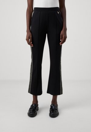 Kobieta ubrana w czarne skrócone spodnie z teksturowanymi bocznymi paskami oraz czarne wsuwane mokasyny, stojąca na jednolitym tle.