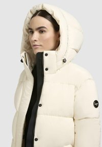 Puffer jacket in cream, featuring a large hood, black zipper, and snap buttons. Textured surface with quilted design and logo patch.