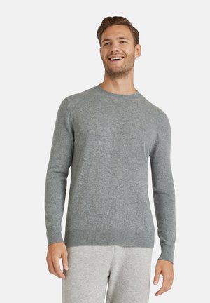 Lächelnder Mann, der einen hellgrauen Pullover mit Rundhalsausschnitt und passende hellgraue Hose trägt, vor einem einfarbigen weißen Hintergrund stehend.