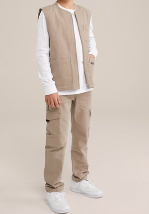 Beige mouwloze vest over een witte blouse met lange mouwen, gecombineerd met beige cargo shorts en witte sneakers. Bevat een borstzak en een rits.