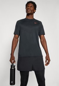 Czarna koszulka sportowa Nike z krótkim rękawem, teksturowana górna część i małe czerwone logo. Model trzyma czarną butelkę na wodę i nosi ciemne szorty.