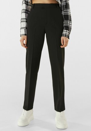 Person wearing black tailored pants paired with white platform sneakers and a black-and-white plaid long-sleeve shirt visible at sleeves.