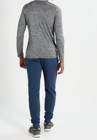 Man wearing gray long-sleeve shirt, blue jogger pants, and green sneakers, standing facing away against a white background.