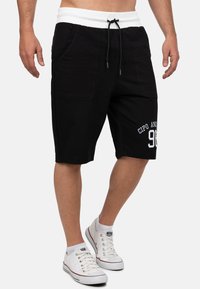 Shorts noirs avec une bande élastique blanche à la taille et un cordon de serrage, comportant deux poches latérales et un motif graphique sur le devant avec des lettres blanches.