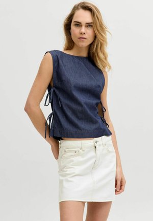 Kvinde med blondt hår iført navyblå ærmeløs top med bindebånd i siden og hvid denimnederdel, poserer med én hånd i lommen mod en ensfarvet baggrund.