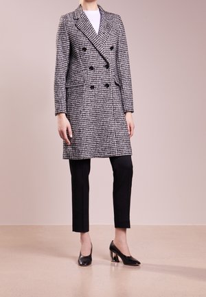 Femme portant un manteau croisé noir et blanc à motif pied-de-poule, un pantalon noir court et des chaussures noires à talons blocs, debout sur un sol beige.