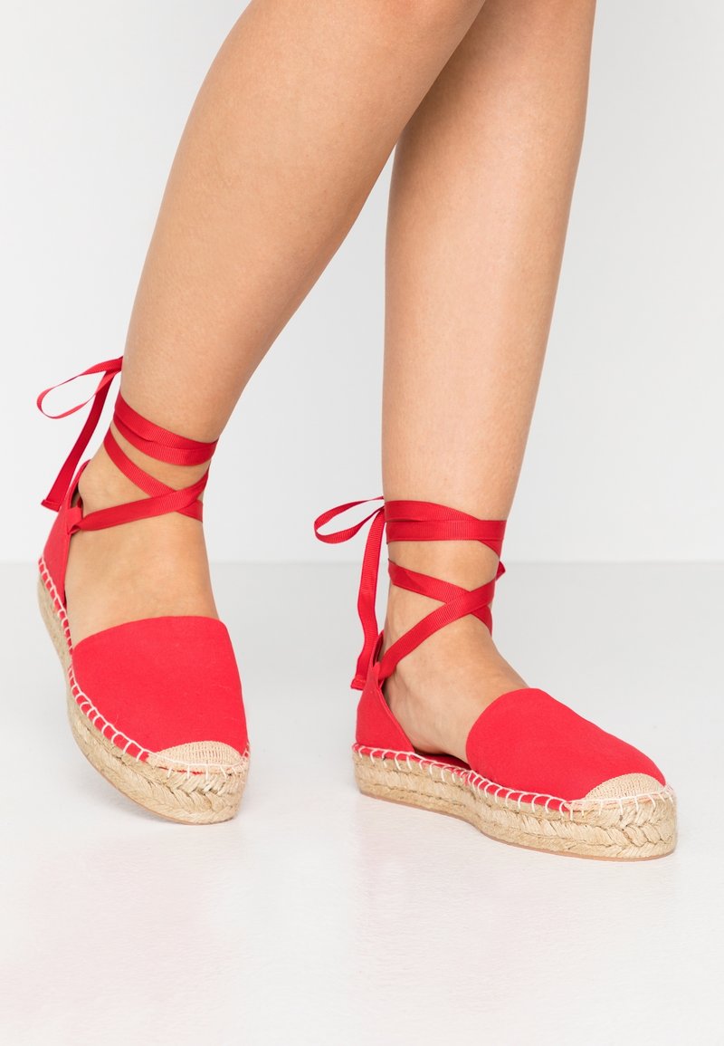 Espadrilles en toile rouge avec une semelle en jute tressé, bout arrondi et rubans autour de la cheville attachés à la jambe. Texture lisse avec des coutures contrastées.
