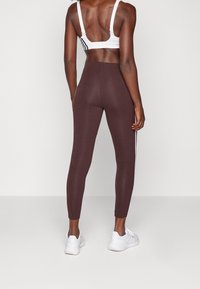 Hnědé legíny s hladkým povrchem a priléhavým střihem, zdobené bílými bočními pruhy. Kombinované s bílou sportovní podprsenkou a bílými teniskami.