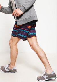 Bañadores de hombre con un diseño multicolor que presenta formas geométricas. Combinados con una sudadera con cremallera gris y zapatillas gris.