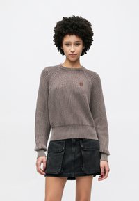 Pruun soonikkoes kampsun, millel on pikad varrukad ja ümar kaelus, ning väike logomärk. Sobitatud musta teksase seelikuga.