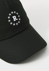 Schwarze Kappe aus strukturiertem Stoff, mit einem weißen gestickten Buchstaben "R" innerhalb eines Kreises von Sternen auf der Vorderseite. Gebogener Schirm.