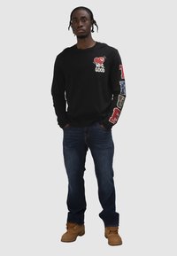 Mann mit geflochtenem Haar, der ein schwarzes Langarmshirt mit rotem Herz und Text, blaue Jeans und beige Stiefel trägt, steht mit den Händen in den Taschen.