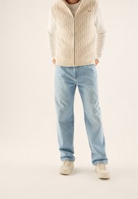 Persona che indossa un gilet in maglia cable-knit color crema, una camicia bianca a maniche lunghe, jeans azzurri chiari e sneakers beige, in piedi con le mani nelle tasche.