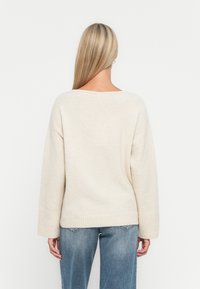 Hellbeige, oversized Strickpullover mit weit ausgeschnittenem Halsausschnitt, gerippten Bündchen und Saum, kombiniert mit hellblauen Jeans.