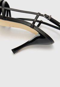 Svart högklackad sandal med tunna korsettsnören och en glansig finish. Har en spetsig tå och en slank stiletto-klack.