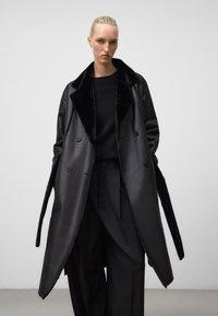 Zwarte dubbelbreasted jas met een satijnen afwerking en een zachte kraag; lange mouwen met een getailleerde pasvorm, gecombineerd met losse zwarte broek.