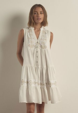 Donna con capelli corti castani che indossa un vestito bianco senza maniche con dettagli beige ricamati e bottoni sul davanti, in piedi su uno sfondo semplice.