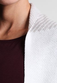 Bílý strukturovaný šál přehozený přes tmavě rubínový top, který má žebrovaný vzor a měkké okraje. Důraz na kontrast teplých barev.