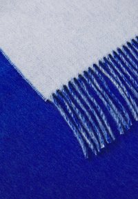 Blå uldplaid med en lysere blå frynsedetail. Har en blød tekstur og et to-tonet farveskema med dyb og lys blå.