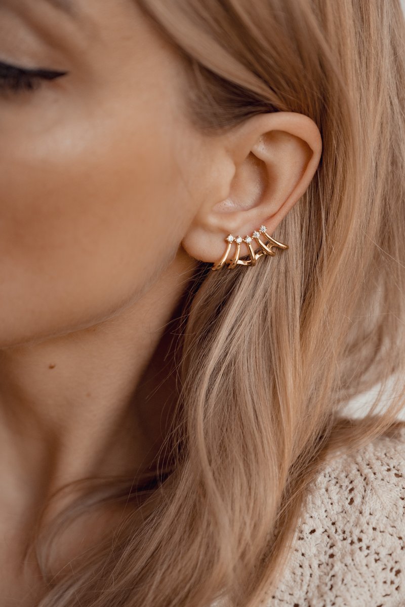 Pendientes de aro dorados que presentan cinco segmentos curvados decorados con pequeñas gemas transparentes, sujetados a la oreja de una persona con el cabello largo y liso.