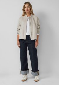 Lichtgrijze bomberjack met ribgebreide manchetten, witte ondershirt, donkere wijde jeans met omslagdetails en witte sneakers op een neutrale achtergrond.