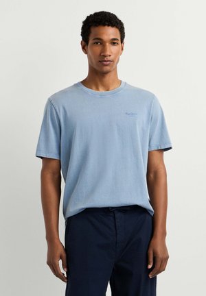 Jonge man met kort krullend haar, gekleed in een lichtblauw t-shirt en donkerblauwe broek, staand tegen een effen achtergrond.
