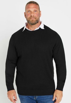 Homme aux cheveux courts et barbe portant un pull noir texturé par-dessus une chemise blanche à col et un jean bleu, debout devant un fond uni.