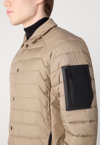 Veste matelassée beige avec un quilting horizontal, une poche zippée noire sur la manche gauche et une fermeture à pression au niveau du col.